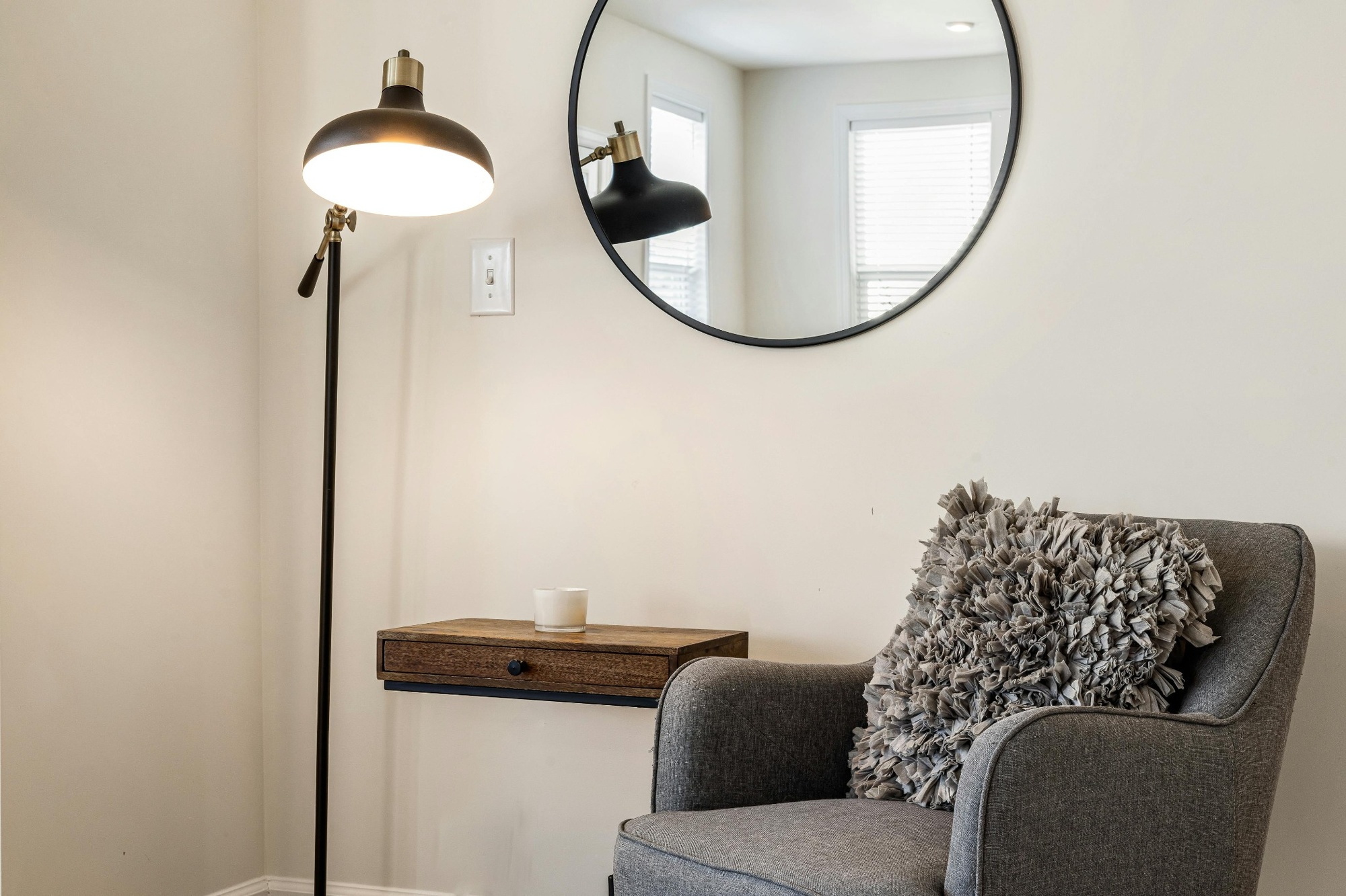 Stock image of a cozy reading corner with a gray armchair textured pillow round mirror lamp and small wall mounted table