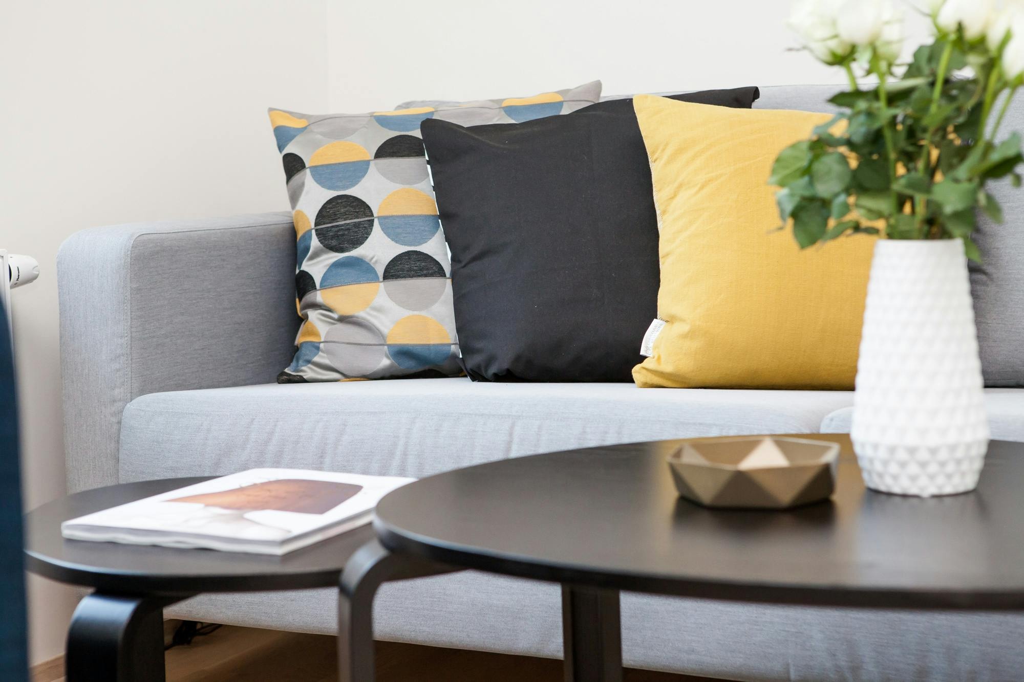 Stock image of a modern sofa with yellow black and geometric pillows beside a white vase of roses