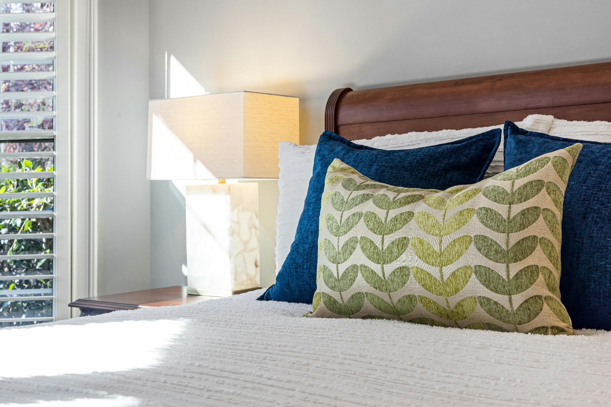 Stock image of a sunlit bedroom with blue pillows green leaf patterned cushion and white bedding